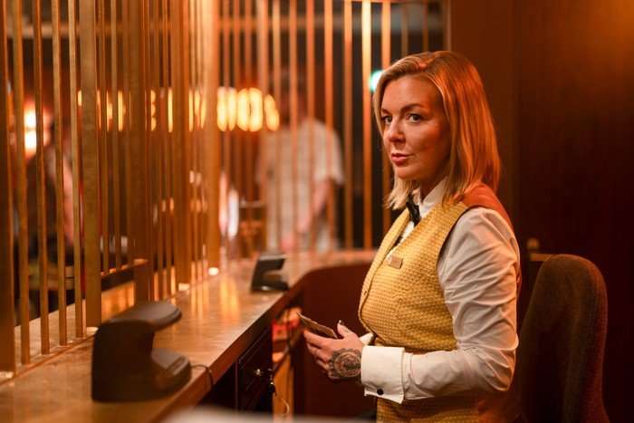Sheridan Smith working in the cage at the casino, wearing a yellow waistcoat and white shirt, counting money and looking to the side.