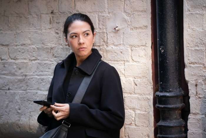 Sophie Mensah standing in an alleyway, wearing a black coat and looking to the side while inspecting her phone.