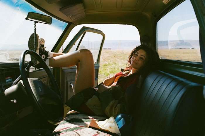 Zendaya laying back in a car, with the seat laid with blankets and resting her feet on the dashboard.