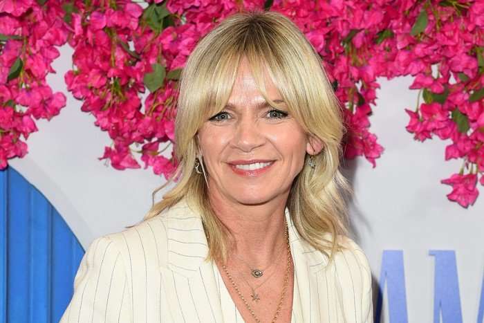 Zoe Ball wearing a white stripy pantsuit, smiling ahead as she poses for a photo. She has straight blonde hair up to her shoulders.