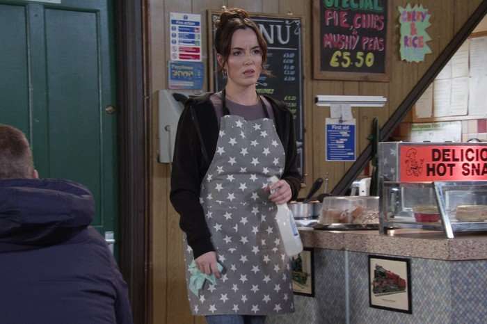 Jodie, in a polkadot apron, holds a bottle of bleach in the Roys Rolls cafe in Coronation Street