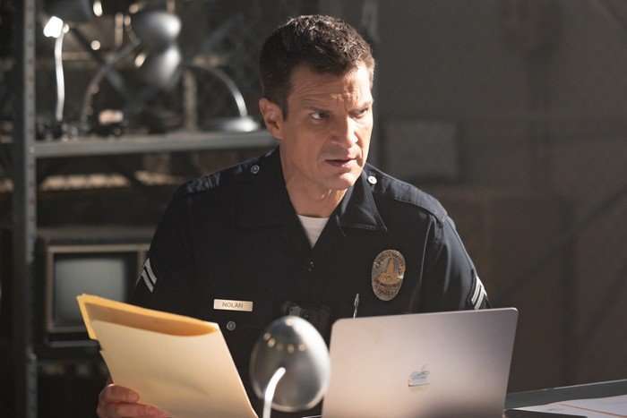 Nathan Fillion in The Rookie, sat at a desk, holding a document