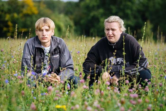 Charlie Cooper as Kurtan and Daisy May Cooper as Kerry in This Country Charlie Cooper as Kurtan and Daisy May Cooper as Kerry in This Country. They are crouched down in a green field. Long grass and wild flowers are sprung up around them.