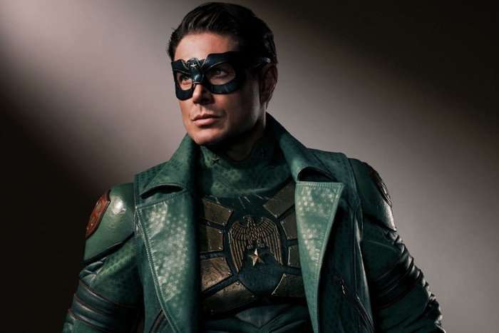 A man in a green superhero outfit, leather jacket and eye-mask poses for the camera