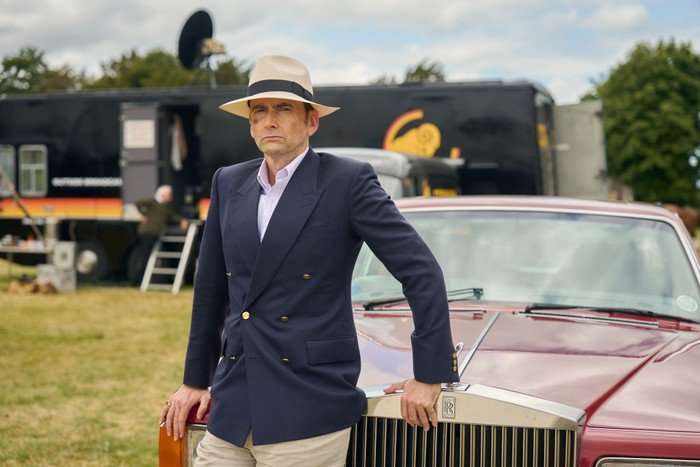 Rivals David Tennant as Tony Baddingham in Rivals, leaning on the hood of a car in a field, wearing a suit and hat.