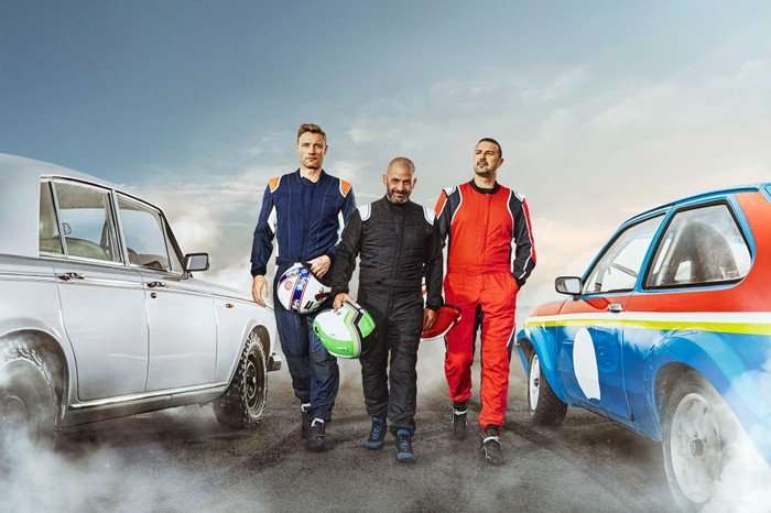 Freddie Flintoff, Chris Harris and Paddy McGuinness for Top Gear. They are wearing driving suits, holding helmets and walking between two cars