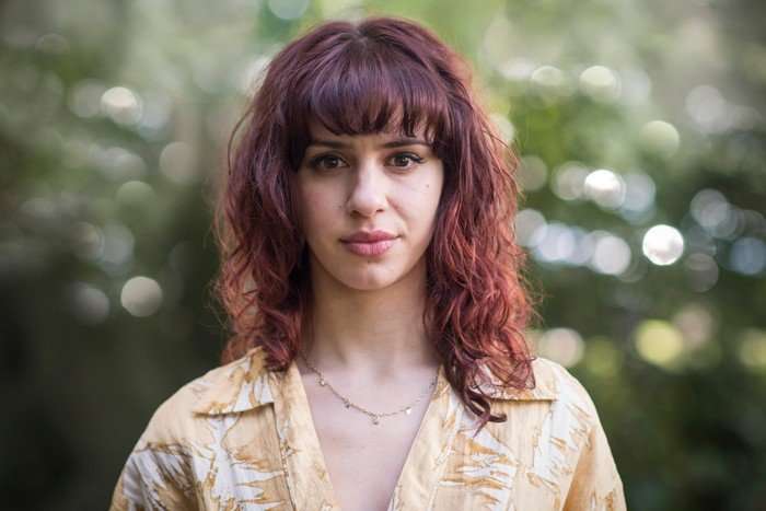 A Good Girl's Guide To Murder A close-up portrait of a woman with shoulder-length wavy auburn-red hair and straight bangs, standing outdoors against a softly blurred green background with circular light bokeh. She is looking directly at the camera with a neutral, calm expression. She’s wearing a light beige patterned button-up shirt and a delicate gold necklace. The natural lighting is soft and even, highlighting her facial features and the warm tones in her hair.