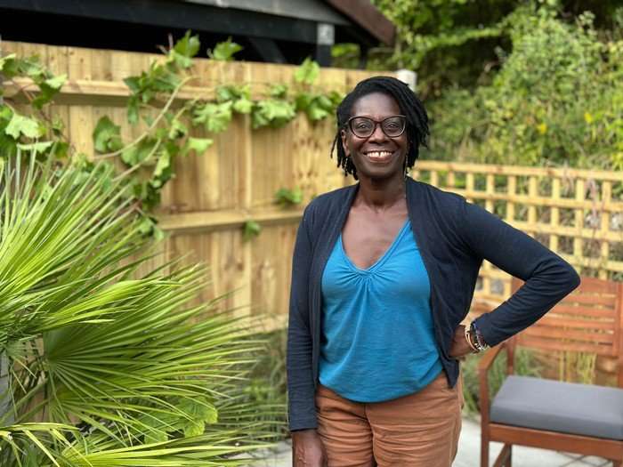 Flo Headlam wearing a blue blouse, brown trousers and a blue cardigan, smiling ahead with one hand resting on her hip.