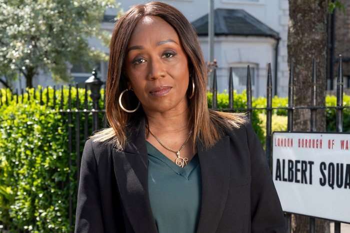Diane Parish as Denise Fox, with her arms folded stood in front of the Albert Square sign in key art for EastEnders