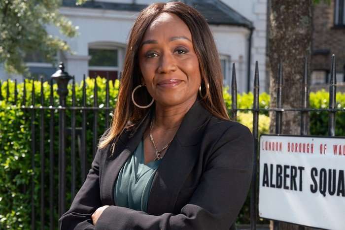 Diane Parish as Denise Fox, with her arms folded stood in front of the Albert Square sign in key art for EastEnders