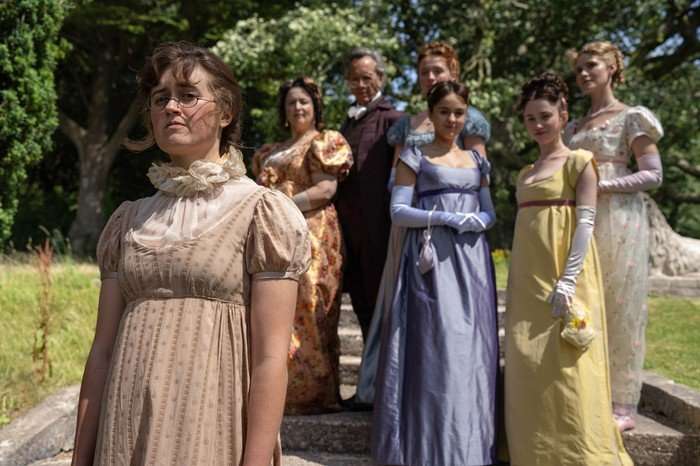 554975,TITLE:The Other Bennet Sister Ella Bruccoleri as Mary Bennet, Ruth Jones as Mrs Bennet, Richard E Grant as Mr Bennet, Poppy Gilbert as Lizzie Bennet, Molly Wright as Kitty Bennet, Grace Hogg-Robinson as Lydia Bennet and Maddie Close as Jane Bennet in The Other Bennet Sister. The women are all wearing dresses and Mr Bennet is wearing a suit, and they are stood outside on some steps. Mary is stood in the foreground, while the others are stood in the background, looking at her.