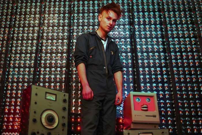 Sam Battle stood next to four boomboxes stacked on top of each other, wearing a blue boiler suit.