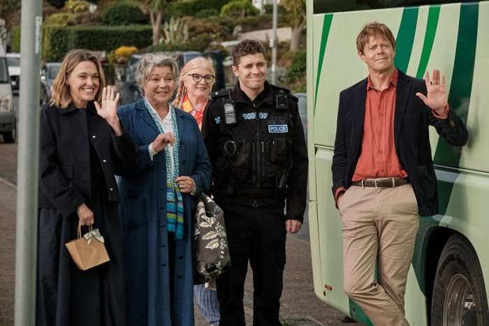 Five people stand on a suburban street beside a green-striped bus, smiling and waving. Three women and a uniformed police officer stand together on the left, while a casually dressed man leans against the bus on the right, also waving. The group appears cheerful, with trees, parked cars, and houses visible in the background.