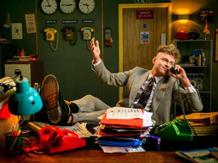 557503,TITLE:Eurovision Song Contest 2026 Sam Battle sat back in a chair with his legs resting on the desk. He is speaking into a phone on his ear with one hand in the air.