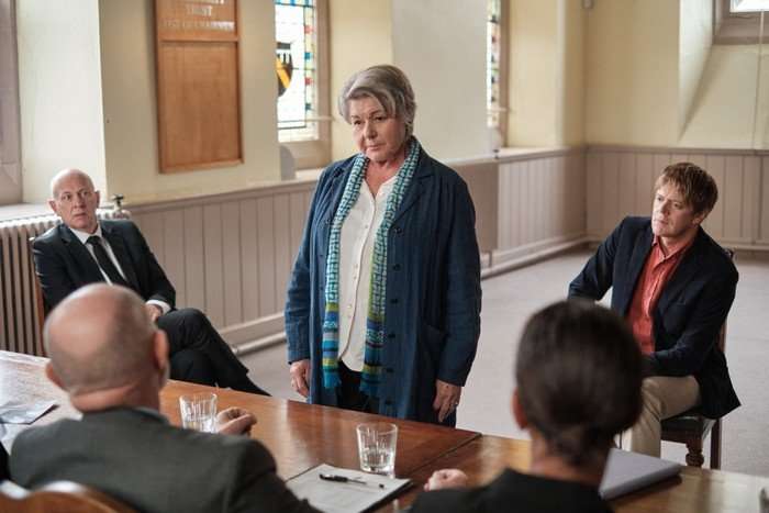 An older woman stands in a modest hall, addressing a small group seated around a table. She wears a blue coat and patterned scarf, her expression serious and resolute. Two men in suits sit behind her, watching, while others are seen from behind in the foreground with notebooks and glasses of water, suggesting a formal meeting or hearing.