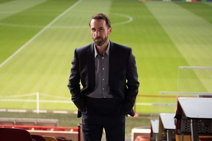 Joseph Fiennes as Gareth Southgate in Dear England, wearing a suit and stood by a football pitch.