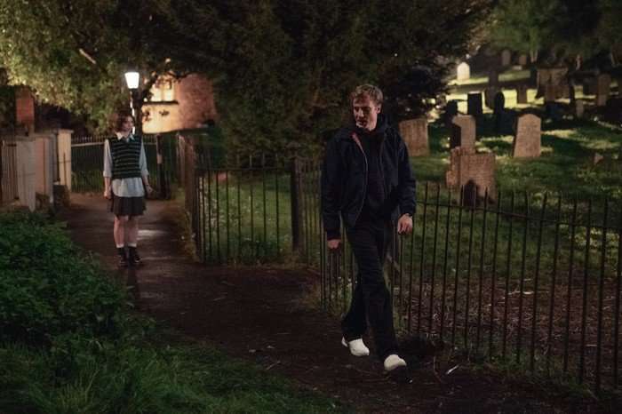Pip stands on a dark pathway beside a graveyard at night while a young man walks ahead of her along the fence line. The scene is lit by streetlamps, with gravestones and trees visible in the background.