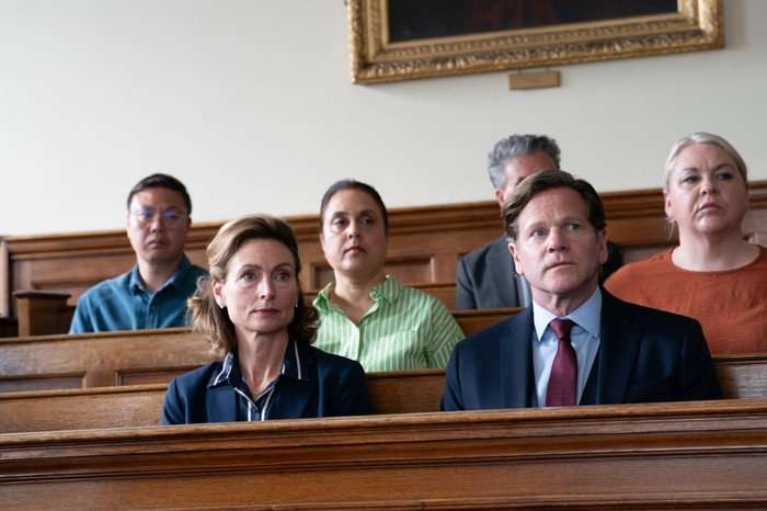 A Good Girl's Guide to Murder series 2 Several people sit on wooden benches inside what appears to be a courtroom or formal hearing room with wood-paneled walls. In the foreground, a well-dressed man in a dark suit and red tie and a woman in a navy blazer sit side by side, both looking ahead with serious, attentive expressions. Behind them are other attendees in casual and business attire, also focused on something out of frame. Soft natural light illuminates the room, and a large framed painting hangs high on the wall above them, adding to the formal atmosphere.