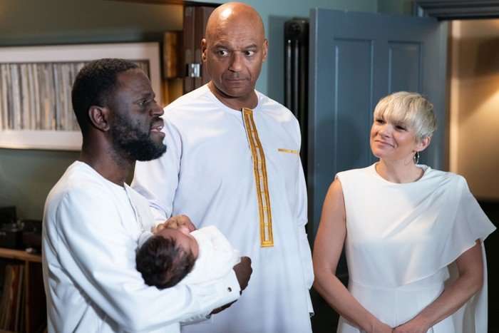 George and Nicola smile towards Kojo, who is holding baby Ivy in a scene from EastEnders