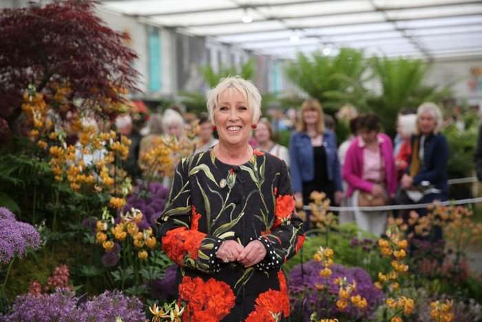 Carol Klein stood inside a garden, smiling ahead and wearing a floral dress.