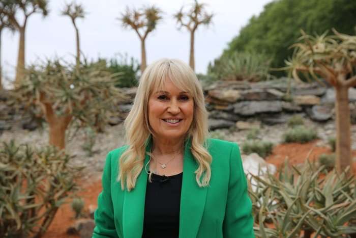 Nicki Chapman wearing a black top and green blazer, smiling ahead as she stands in a garden.