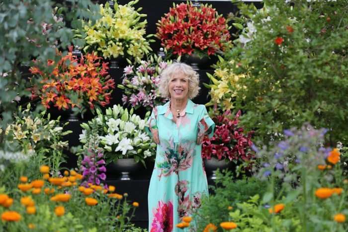 Sue Kent wearing a light blue dress with floral prints, stood around a flower garden.
