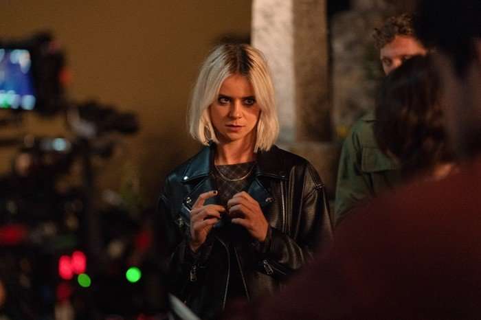 A Good Girl's Guide to Murder series 2 A young woman with a short platinum-blonde bob haircut stands indoors in a dimly lit setting, wearing a black leather jacket over a dark top. She has an intense, focused expression and appears to be engaged in conversation or observing something off-camera. Warm orange lighting casts dramatic shadows across the scene. In the blurred background, other people are partially visible, along with filmmaking equipment and small colored lights, giving the image a behind-the-scenes or nightlife atmosphere.