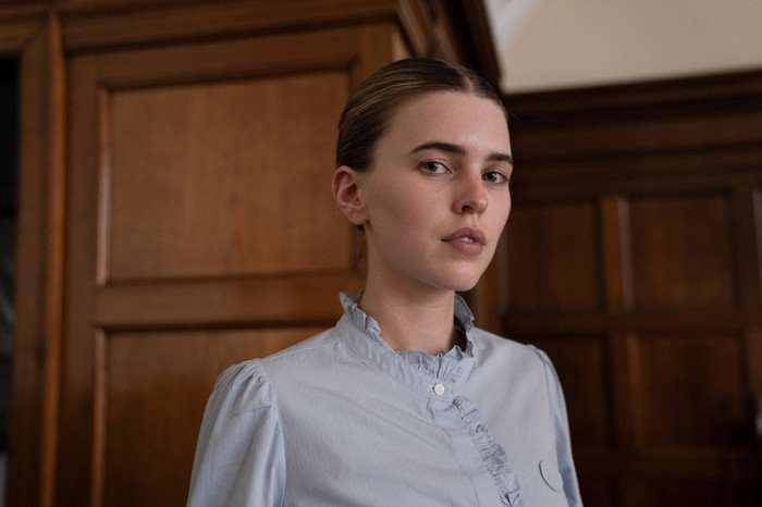 A Good Girl's Guide to Murder series 2 A portrait of a young woman standing indoors in front of dark wooden paneling and doors. She has her hair neatly pulled back and is wearing a pale blue blouse with ruffled trim along the collar and front. She looks toward the camera with a composed, slightly serious expression. The lighting is soft and natural, giving the image a calm, refined atmosphere with warm wood tones contrasting against her light-colored clothing.