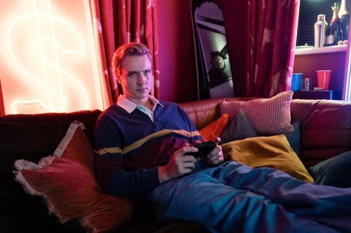 A Good Girl's Guide to Murder series 2 A young man lounges on a couch in a dimly lit room illuminated by colorful neon lighting. He’s holding a video game controller and looking directly toward the camera with a serious, focused expression. He wears a long-sleeved rugby-style shirt with horizontal stripes and loose blue jeans. The room has a cozy, nightlife atmosphere with red curtains, decorative pillows, bottles on a shelf, and a glowing neon dollar-sign light on the wall behind him.