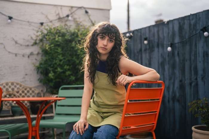 A Good Girl's Guide to Murder series 2 A young woman with long, dark curly hair and bangs sits outdoors on a bright orange metal chair in a small patio or garden area. She’s wearing a sleeveless olive-green dress layered over blue jeans. One arm rests casually on the chair back while she looks directly at the camera with a calm, thoughtful expression. Around her are matching orange and green patio furniture, string lights, potted plants, and a dark wooden fence. The lighting is soft and natural, with sunlight creating a warm glow around her hair and shoulders.
