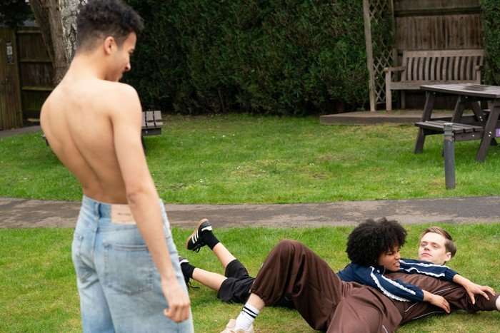 Josh stands shirtless over Jasmine and Oscar, who are lying on some grass, in EastEnders