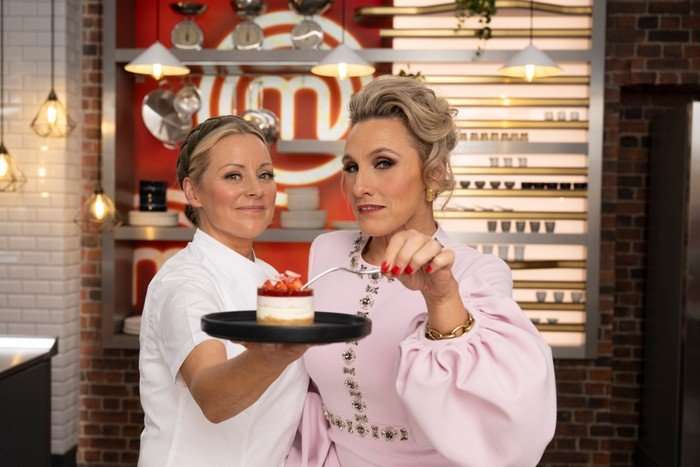 Anna Haugh and Grace Dent stood next to each other, smiling ahead. Anna is holding a plate with a cheesecake on it as Grace leans forward with a fork.
