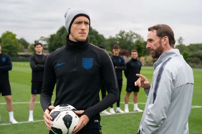 561323,TITLE:Dear England Will Antenbring as Harry Kane and Joseph Fiennes as Gareth Southgate in Dear England. Kane is holding a football, wearing black and stood in front of a line of other players. Southgate is stood next to him wearing a grey jacket.