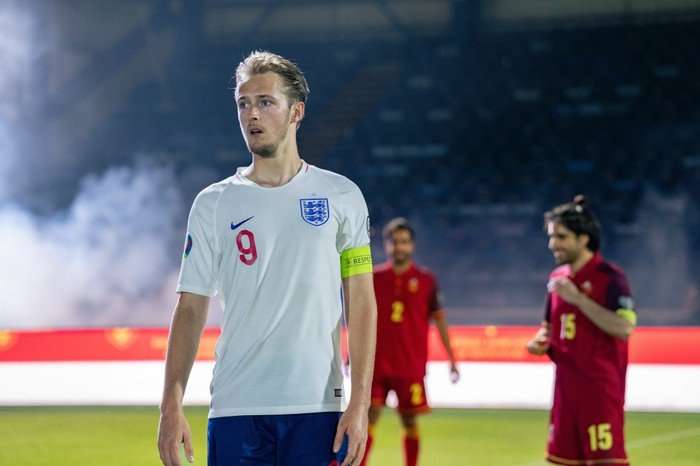 Will Antenbring as Harry Kane in Dear England, with a white England football kit and stood on the pitch, with players in red stood behind him.