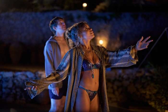 Antonia Thomas as Jess and Damien Molony as Dan in Two Weeks in August, stood together outside at night. They are both in swimming costumes, she is wearing a robe on top while he is wearing a jacket.