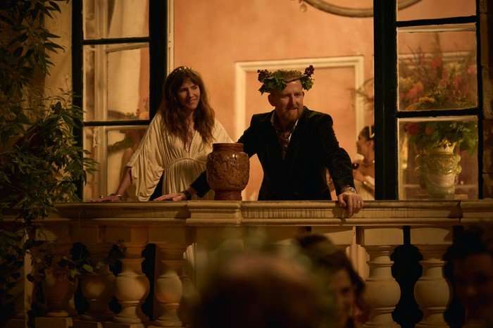 561503,TITLE:Two Weeks In August Dolly Wells as Flick and Tom Goodman-Hill as James in Two Weeks in August, wearing costumes and stood on a balcony together at night.