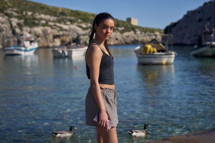 561623,TITLE:Two Weeks In August Maria Almeida as Avery in Two Weeks in August, stood by a body os water, wearing grey shorts and a black top and holding her phone.