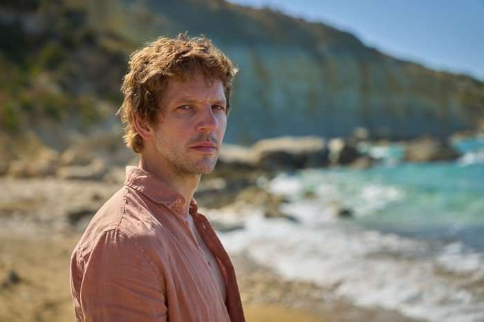 561638,TITLE:Two Weeks In August Damien Molony as Dan in Two Weeks in August, wearing a pink shirt, stood on a beach and looking at the camera.
