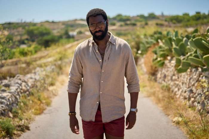 561641,TITLE:Two Weeks In August Nicholas Pinnock as Solomon in Two Weeks in August, wearing a brown shirt, red shorts and sunglasses, walking along a road.
