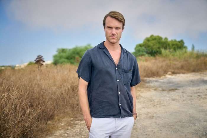 561642,TITLE:Two Weeks In August Hugh Skinner as Jacob in Two Weeks in August, wearing a blue shirt and white shorts, stood on a road.