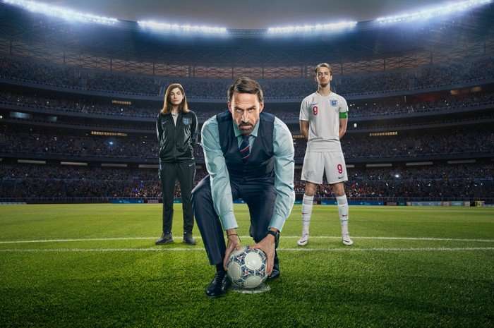 Jodie Whittaker as Pippa Grange, Joseph Fiennes as Gareth Southgate and Will Antenbring as Harry Kane in Dear England. They are stood in a football stadium on the pitch. Gareth is wearing a shirt, tie and waistcoat, and is leaning down to hold a football. Pippa is stood behind one side wearing a tracksuit, and Harry is stood the other side wearing a football kit.