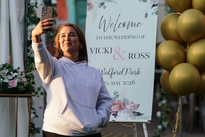 Vicki, in a grey hoody, takes a picture in front of a sign welcoming guests to her wedding with Ross in a scene from EastEnders