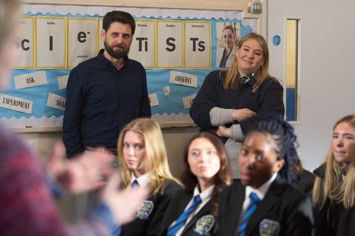 A man with a beard and a blonde woman stand at the back of a classroom, watching attentively as students sit in front of them. The students are wearing black school uniforms with blue striped ties. Behind the adults is a colorful classroom display board styled like a periodic table, with large letters spelling part of the word &ldquo;scientists.&rdquo; Various educational signs such as &ldquo;Ask,&rdquo; &ldquo;Observe,&rdquo; and &ldquo;Experiment&rdquo; are pinned around the board. The atmosphere suggests a school presentation or assembly in progress.
