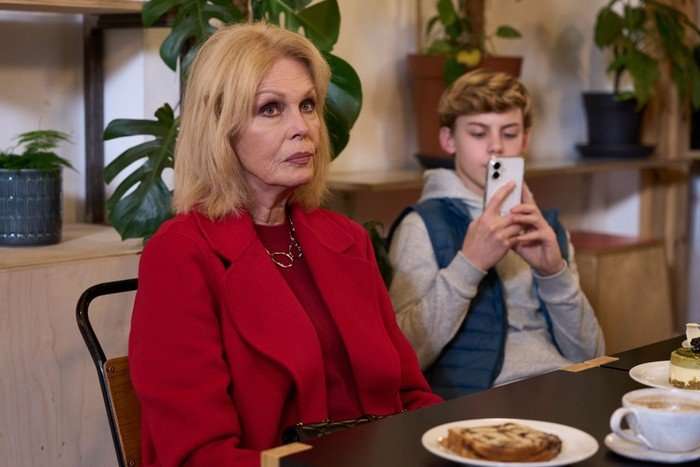 A blonde woman wearing a bright red coat and matching top sits at a caf&eacute; table with a serious, thoughtful expression. She is seated in the foreground beside plates of pastries and a cup of coffee. Behind her, a teenage boy in a gray hoodie and blue vest is focused on his smartphone, holding it up as if taking a photo or recording. The setting is warm and modern, with large green houseplants and wooden shelving in the background.