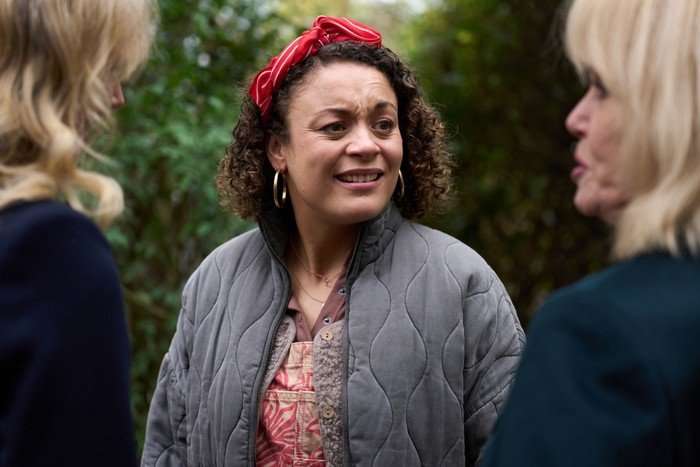 A woman with curly hair and a red striped headband stands outdoors between two other women who are partially out of focus in the foreground. She is wearing large hoop earrings, a gray quilted jacket, and layered casual clothing with a patterned top underneath. Her expression appears concerned or emotional as she speaks. The background is filled with greenery and trees, suggesting a park or garden setting.