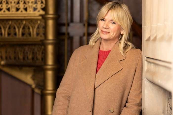 562217,TITLE:Who Do You Think You Are? s23 Zoe Ball stood resting her body to one side. wearing a red jumper and light brown jacket.