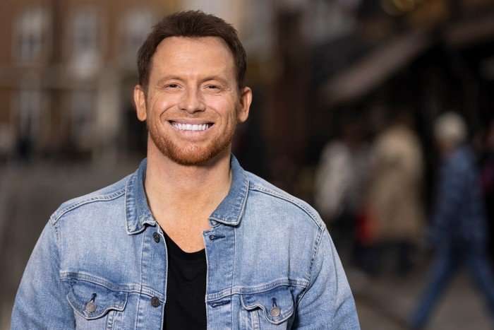 562220,TITLE:Who Do You Think You Are? s23 Joe Swash stood smiling ahead, wearing a black t-shirt and blue denim jacket.