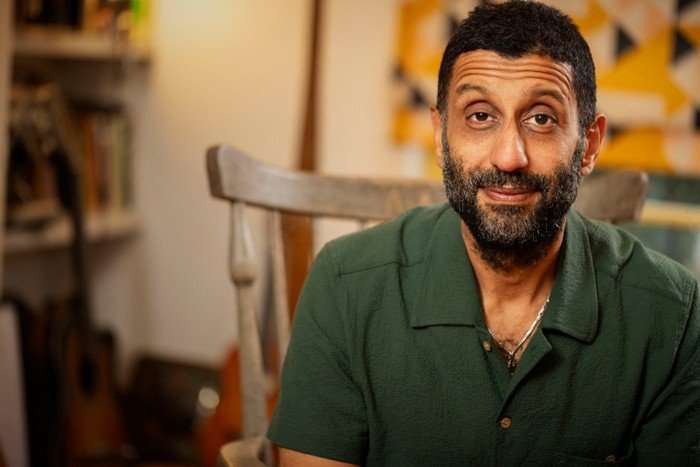 562224,TITLE:Who Do You Think You Are? s23 Adeel Akhtar sat on a chair, smiling ahead, as he wears a dark green shirt. He has black hair and a beard with grey colours.