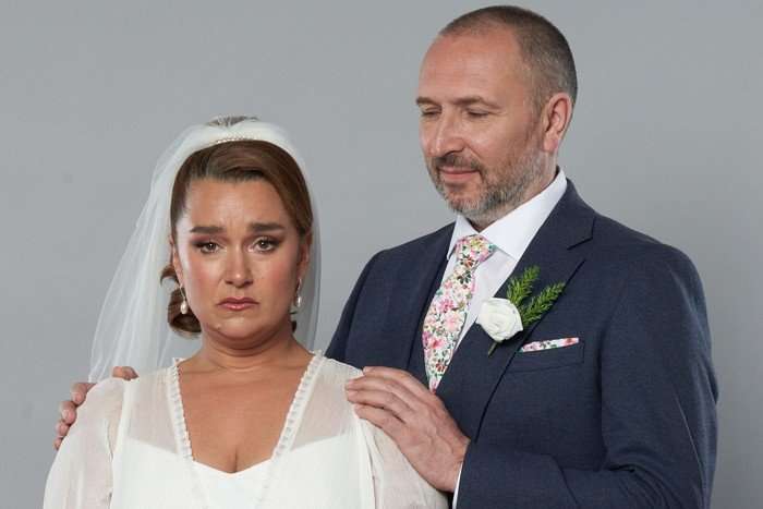 Ross, in a suit, puts his hand on Vicki's shoulder in key art for EastEnders. She is wearing a white wedding dress and veil.
