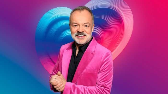 Graham Norton wearing a black shirt, crushed pink velvet blazer stood smiling ahead with his hands placed together.